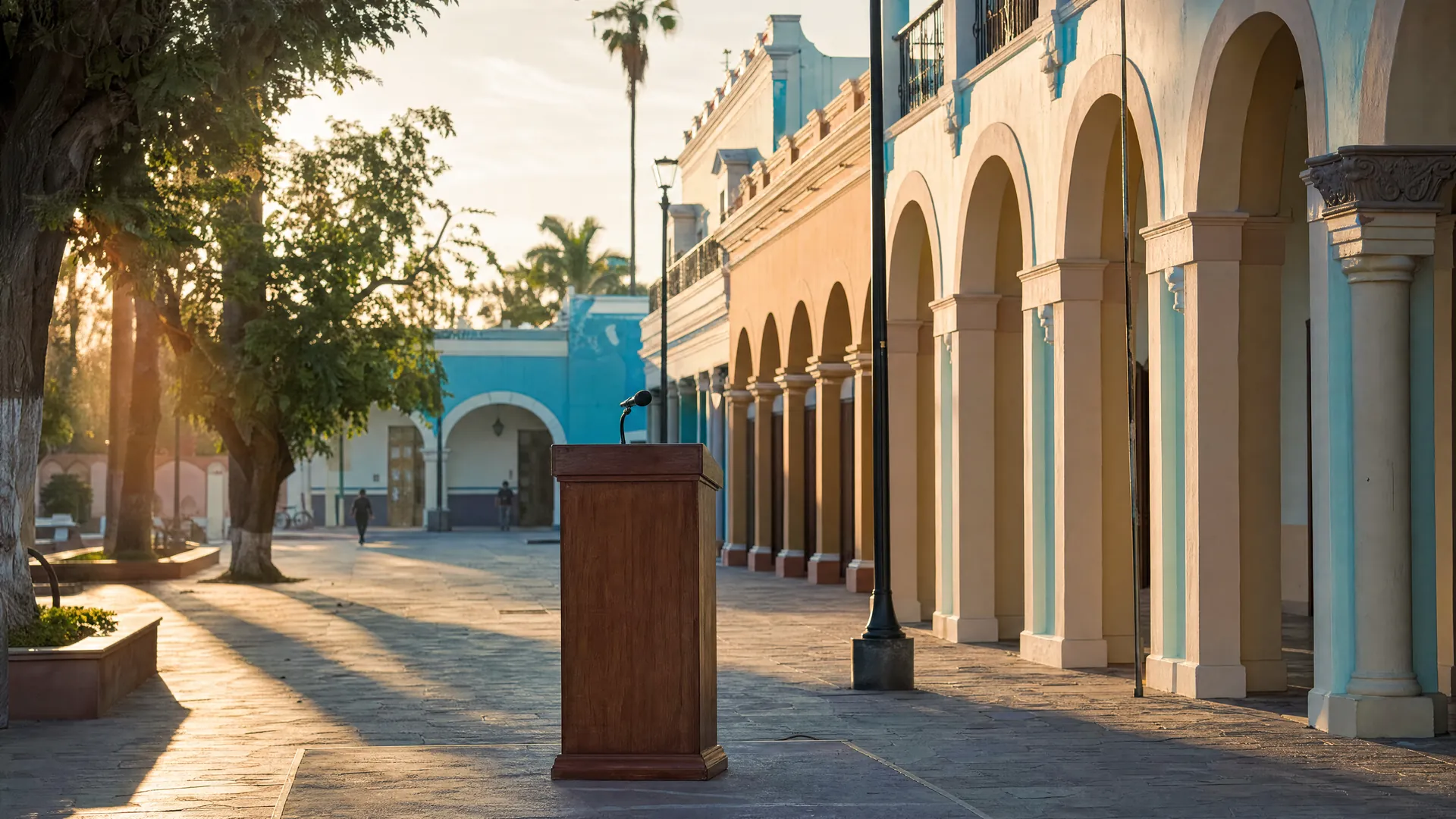 Podio vacío con micrófono en una plaza colonial de La Paz, con arcos de estuco pastel y palmeras bajo la luz dorada de la mañana.