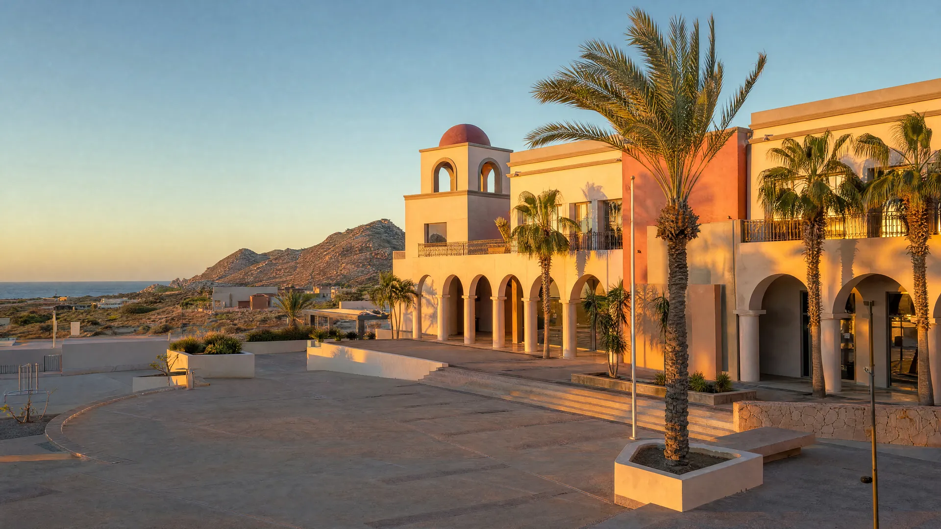 Edificio de gobierno municipal de fachada pastel con arcos y palmeras en Los Cabos al atardecer, con cerros desérticos y el mar al fondo.