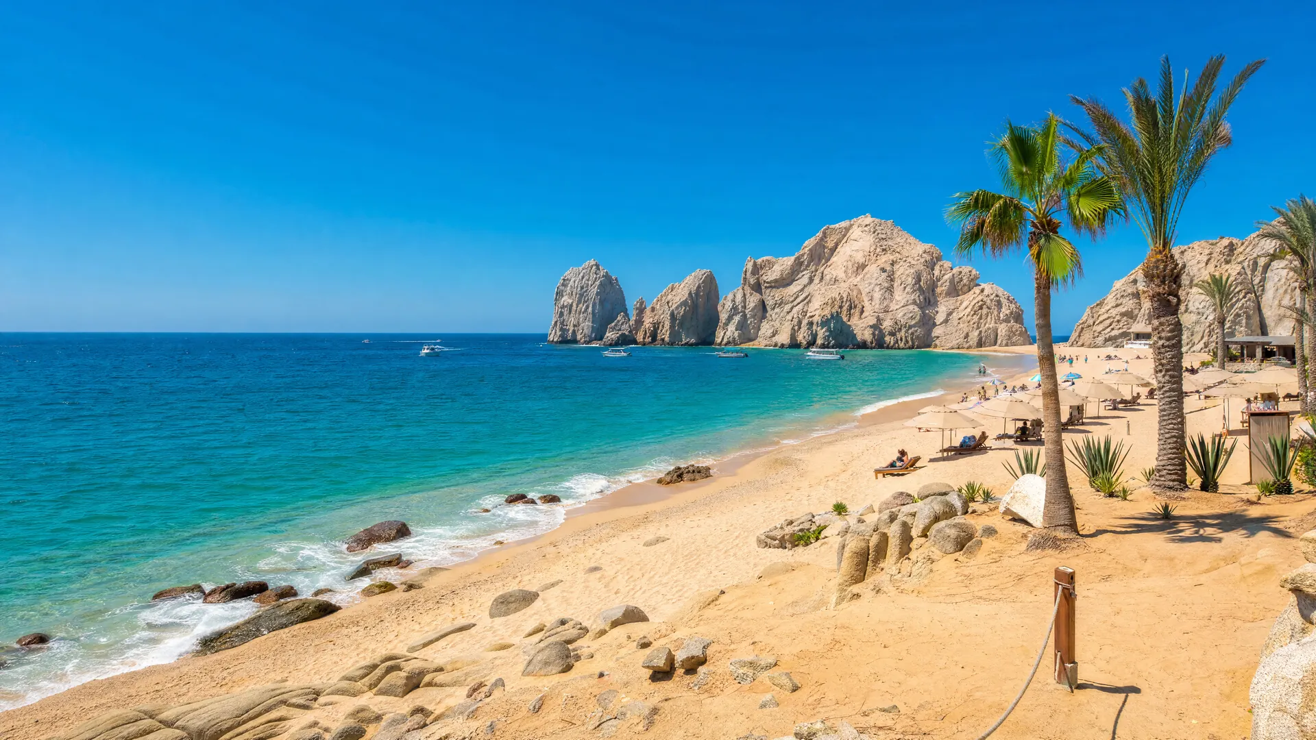 Bahía de Cabo San Lucas bajo cielo despejado y sol intenso, con arcos de roca, palmeras y figuras lejanas bajo sombrillas en la playa.