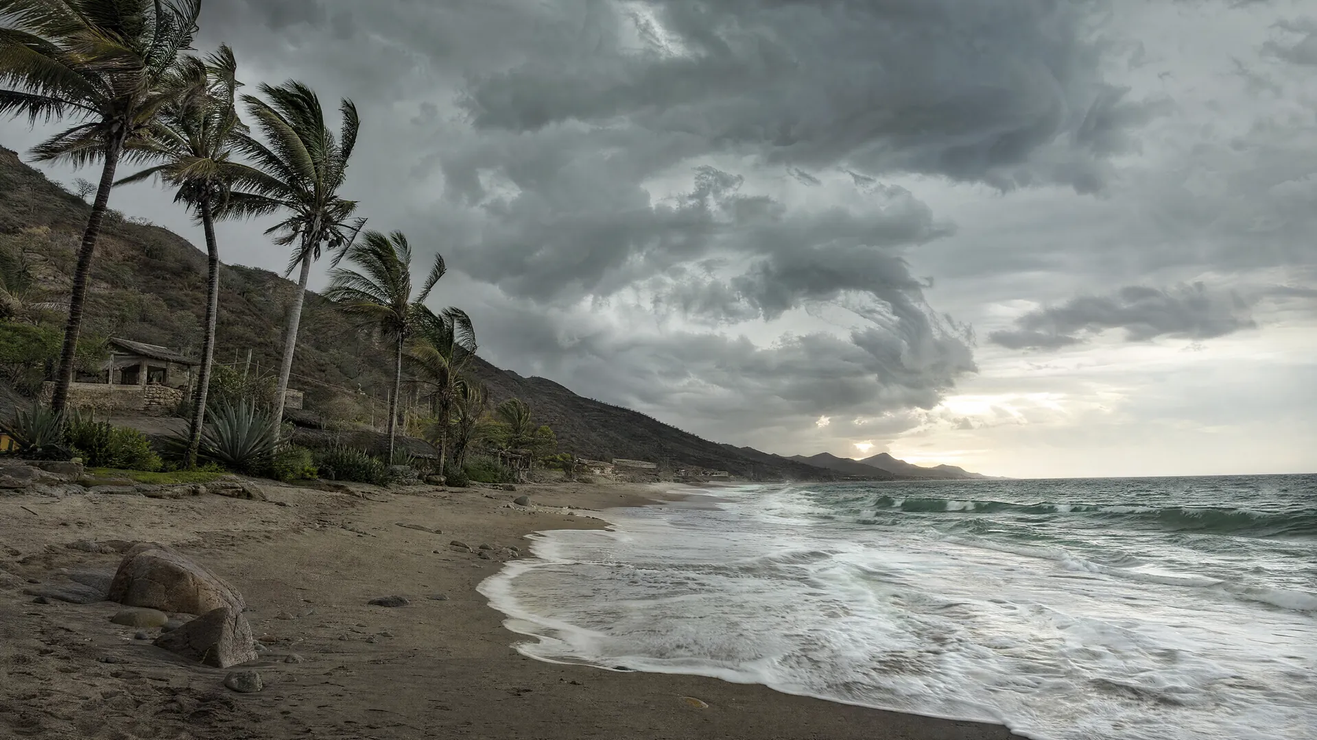 Costa del Pacífico de Baja California Sur con cielo nublado de tormenta, oleaje sobre la playa vacía y palmeras movidas por el viento