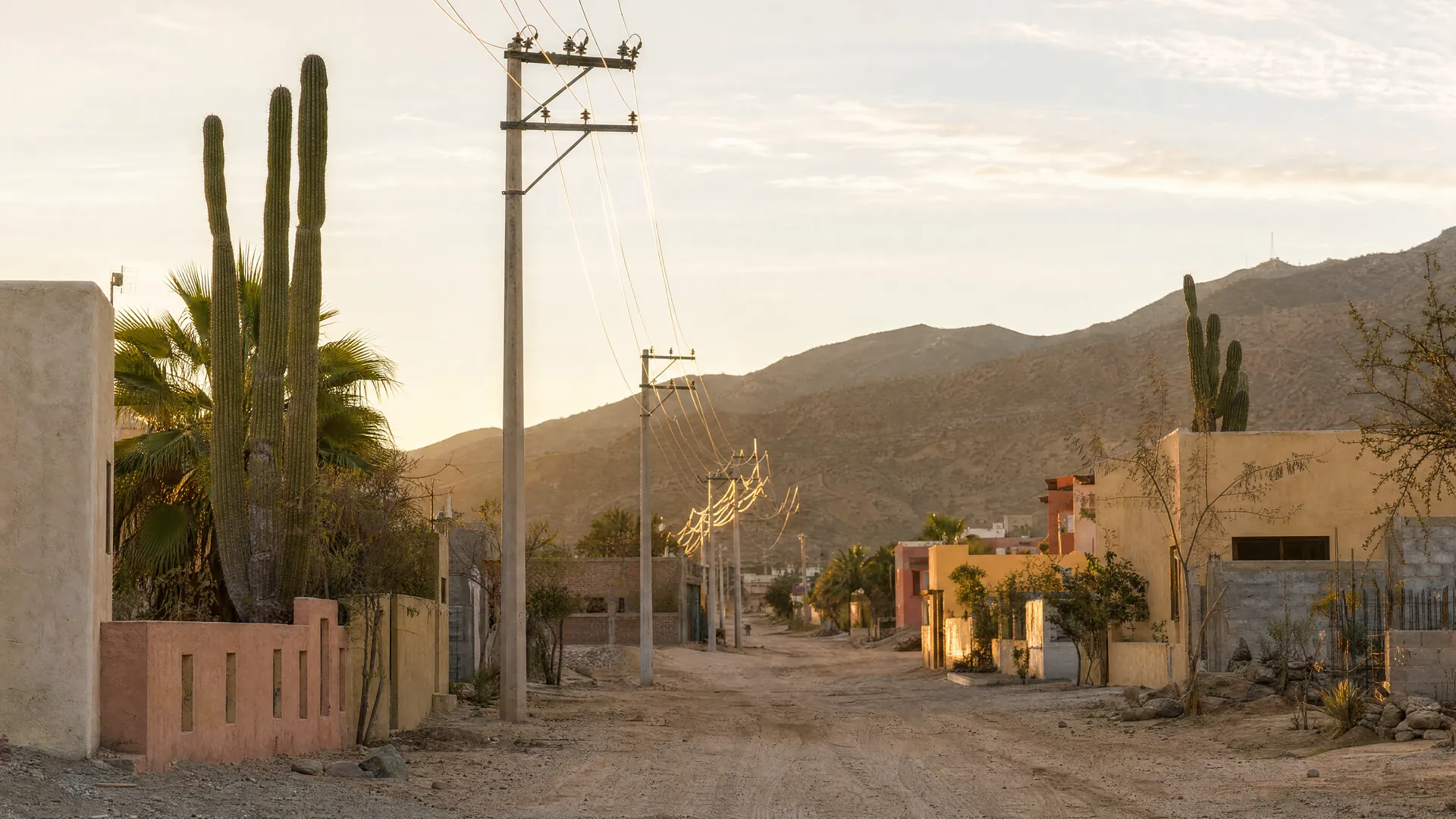 Postes de luz nuevos con cables a lo largo de un camino de tierra en una colonia desértica de Los Cabos, con casas de colores al atardecer