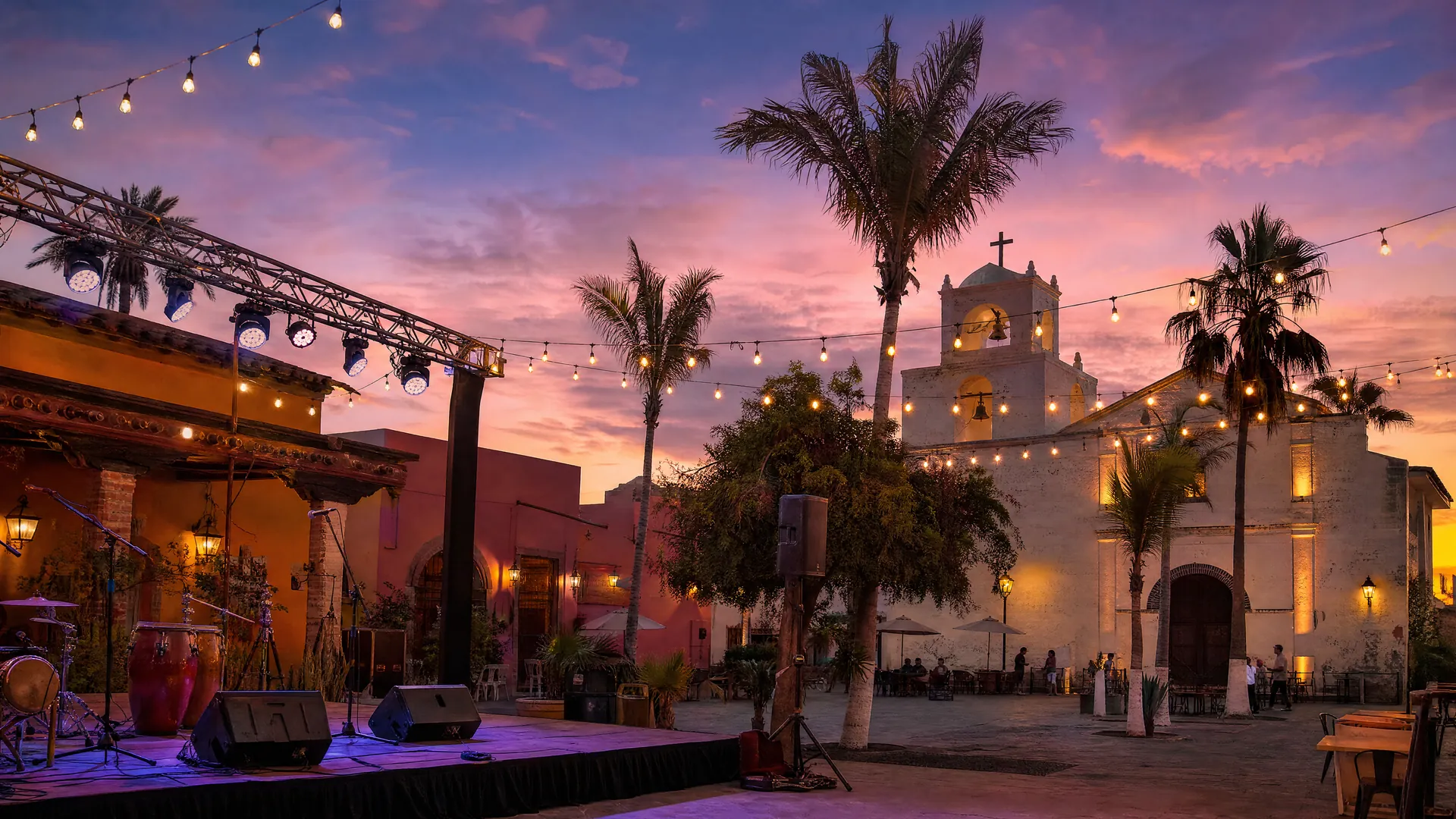 Escenario vacío con instrumentos y luces colgantes en la plaza de Todos Santos al atardecer, con iglesia, palmeras y edificios de colores.