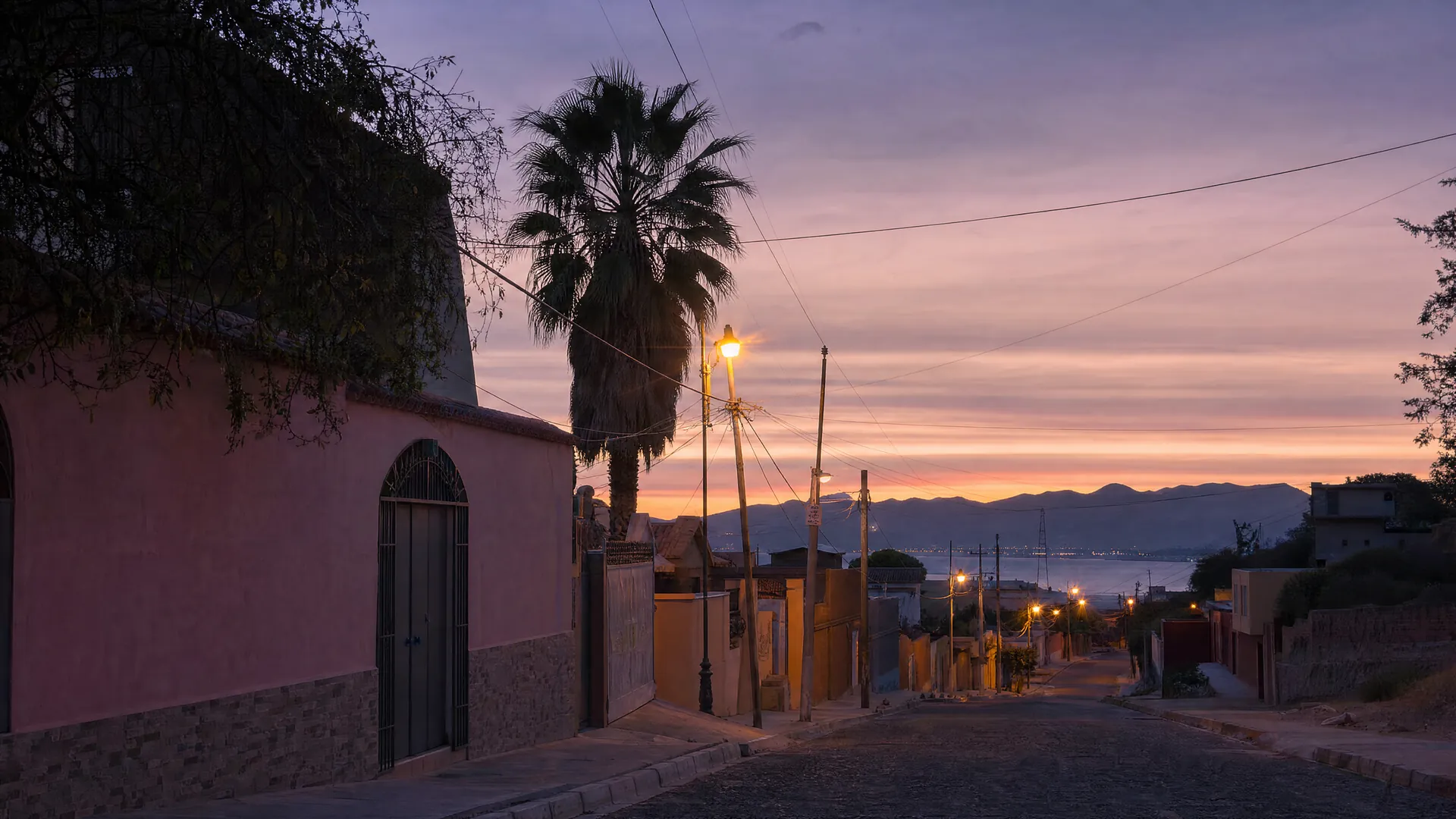 Calle residencial tranquila en La Paz al anochecer, con casas de estuco en tonos pastel, una palmera y faroles encendidos, sin personas.