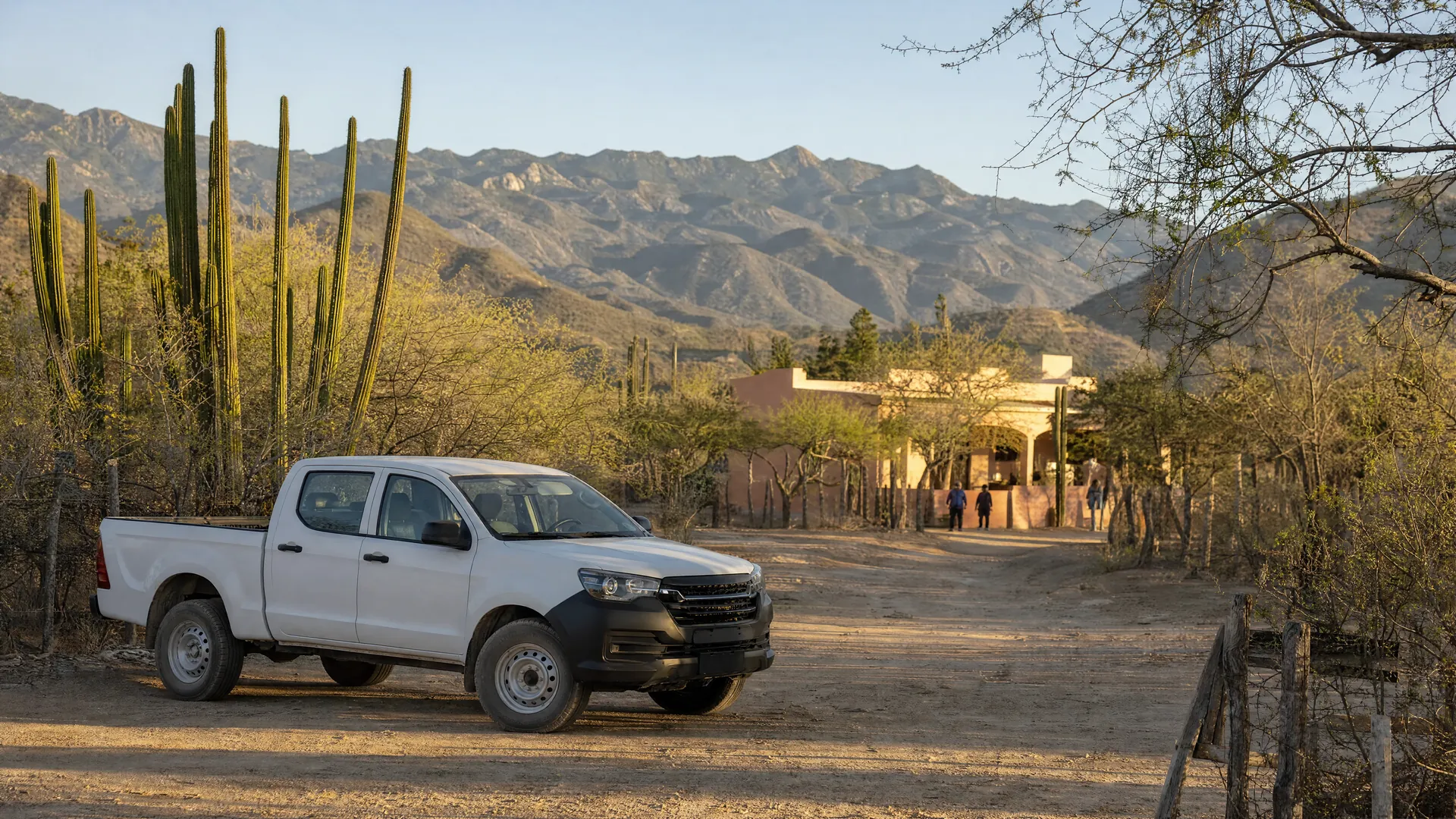Camioneta de patrulla estacionada en un camino de terracería junto a una comunidad rural con cactus y sierra al fondo, al amanecer.